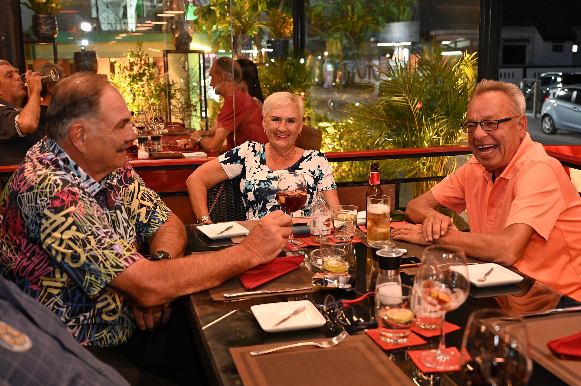 DSC 7089 Three friends laugh and toast over drinks during a lively evening dinner at a cozy plant-filled restaurant